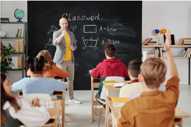 A Teacher facing rows of students, one student raises their hand